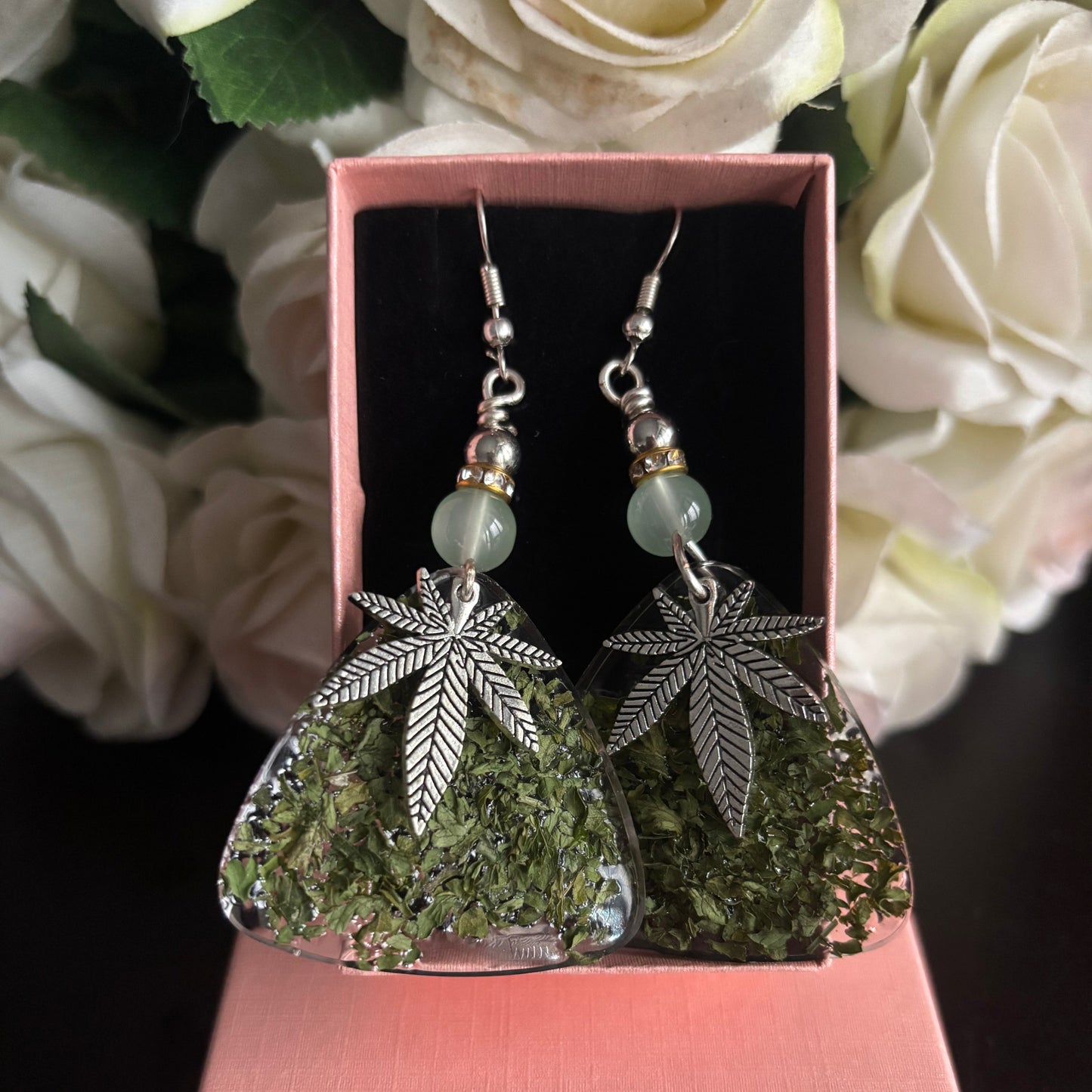 A pair of dangling earrings displayed in an open pink box, with dried green leaves inside clear triangular pendants and silver-tone leaf charms, set against a background of white roses.