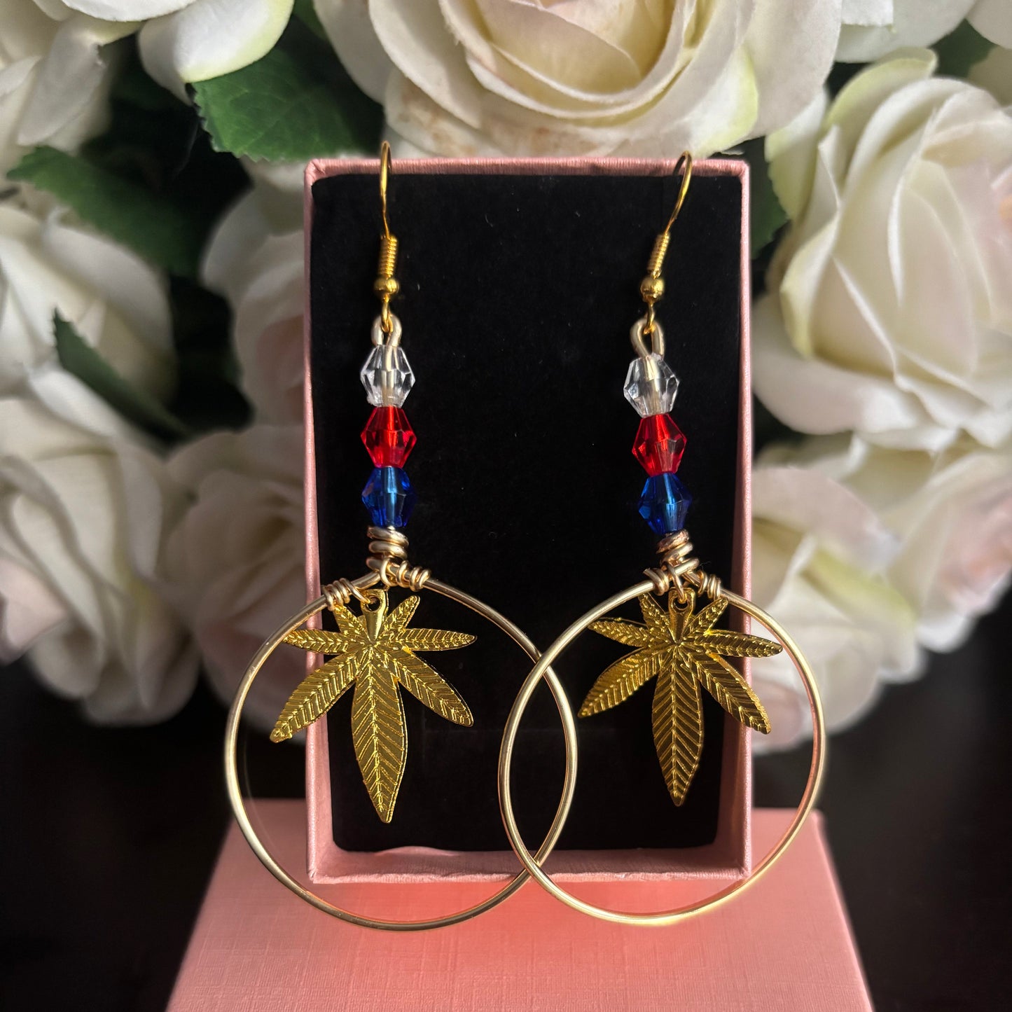 A pair of gold hoop earrings with dangling gold leaf charms, red and blue beads, and clear crystal accents, displayed on a pink jewelry box with white flowers in the background. Show your American Pride, Puerto Rican Pride or 420 pride!