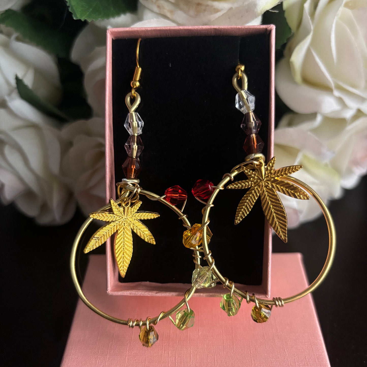 A pair of gold hoop earrings with dangling maple leaf charms, red beads, and clear crystal accents, displayed in a pink jewelry box with white roses in the background.  Celebrate fall with autumn colors and your favorite 420 leaves!