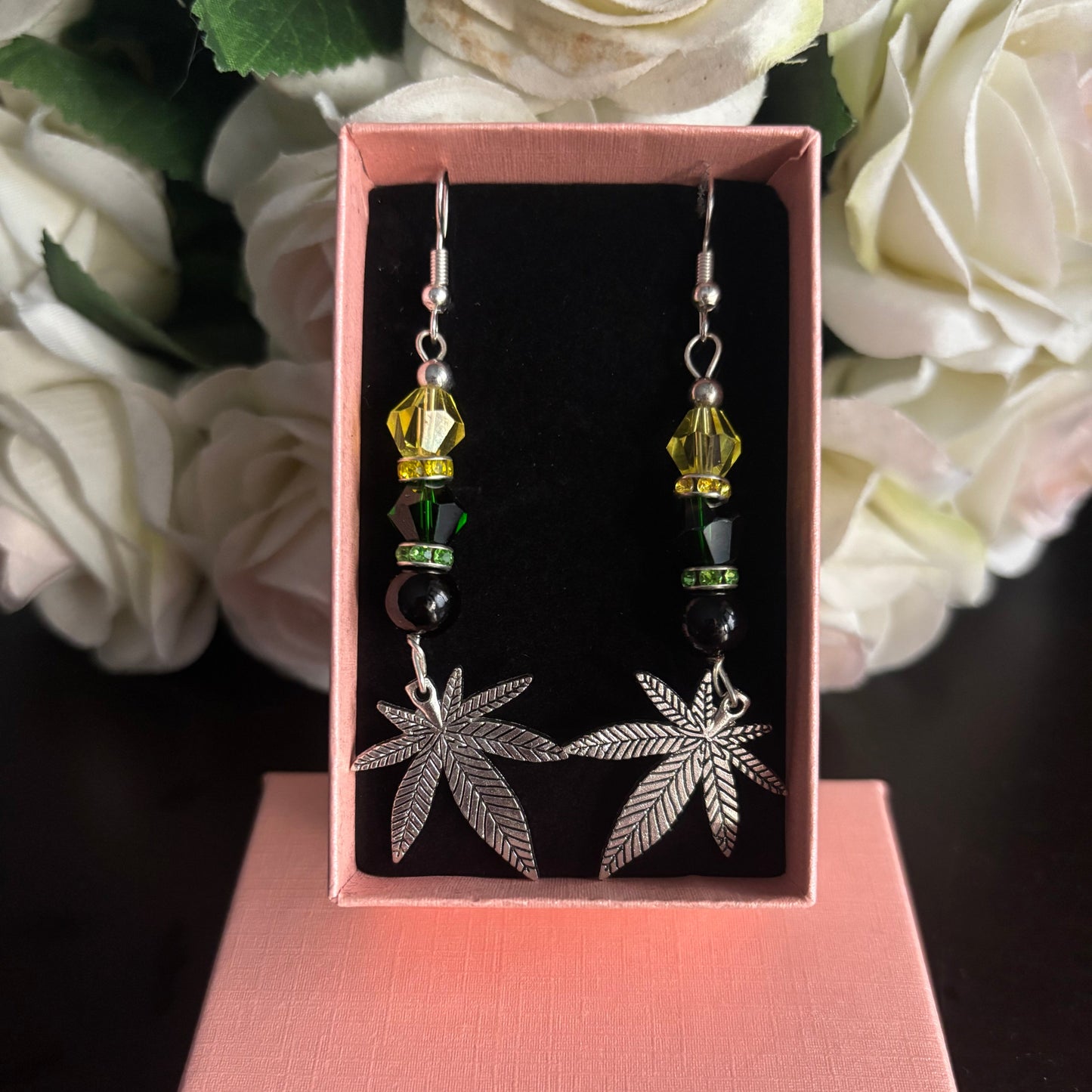 A pair of dangling earrings displayed in an open pink gift box. Each earring has a silver-tone leaf charm, a yellow faceted bead, a dark green bead, and a black bead, with a silver fishhook at the top.