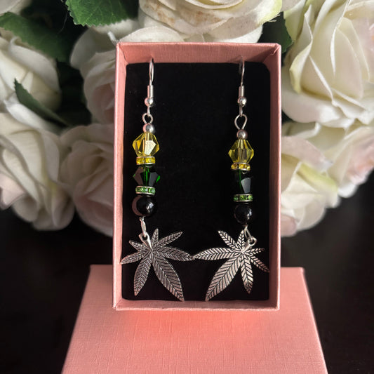 A pair of dangling earrings displayed in an open pink gift box. Each earring has a silver-tone leaf charm, a yellow faceted bead, a dark green bead, and a black bead, with a silver fishhook at the top.