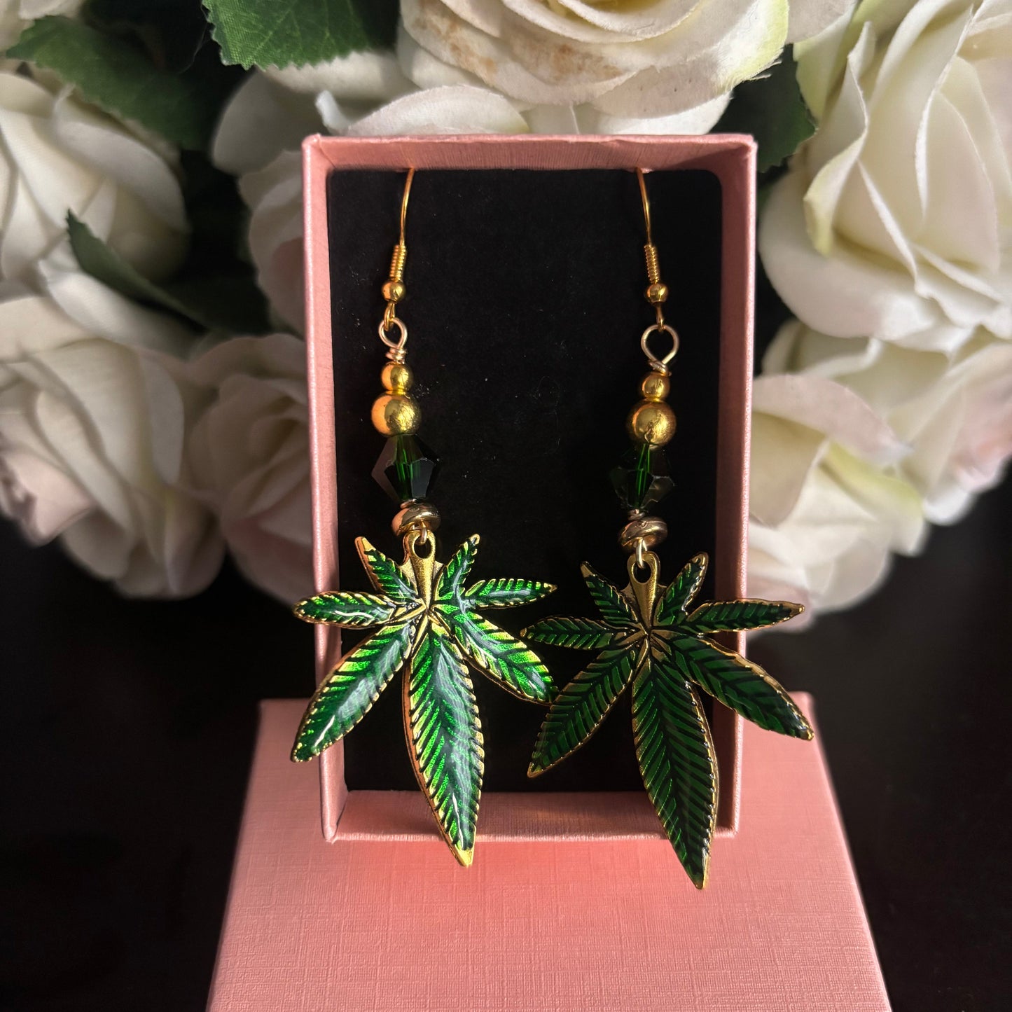 A pair of dangling earrings shaped like green cannabis leaves with gold-tone outlines and small gold beads above, displayed in a pink jewelry box with white roses in the background.