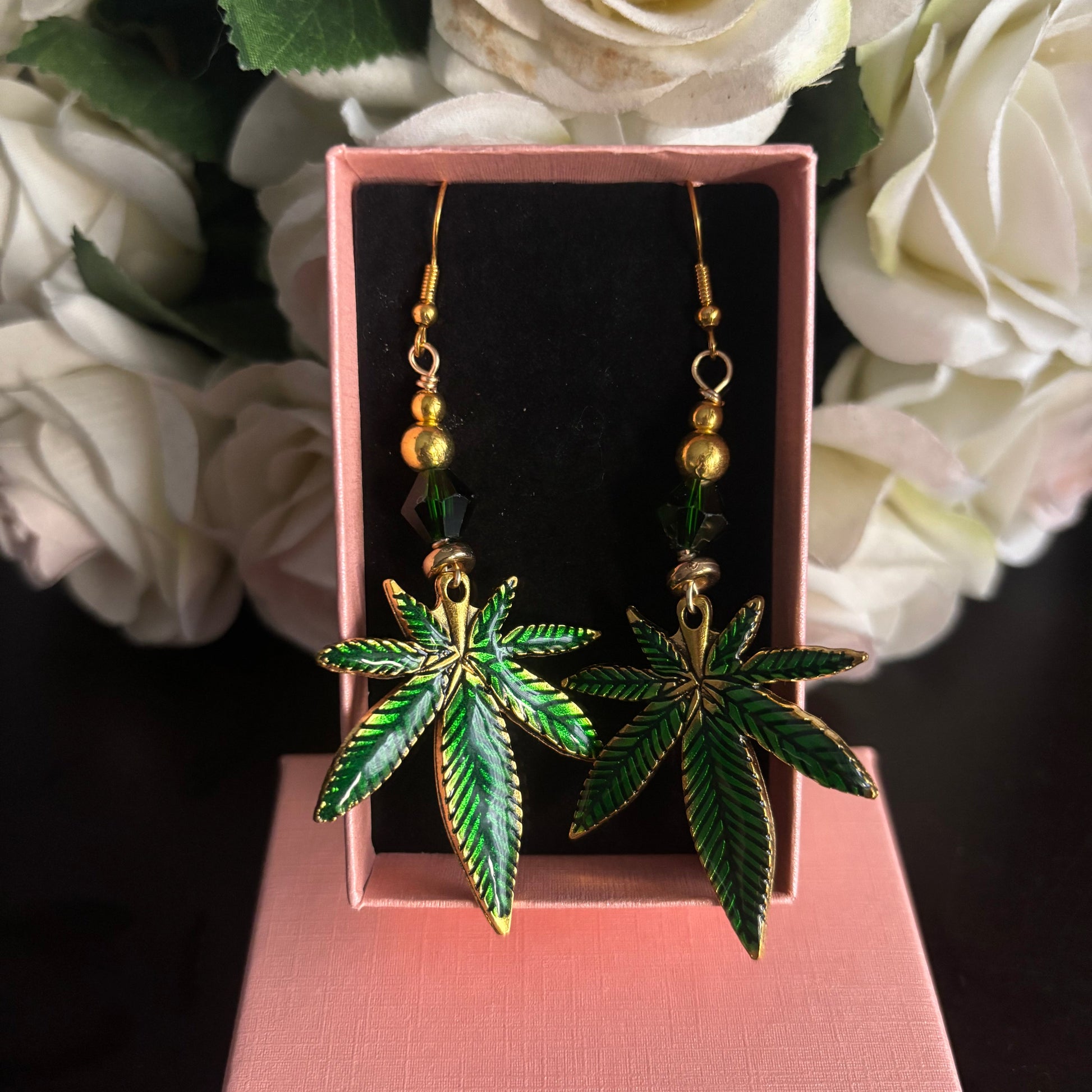 A pair of dangling earrings shaped like green cannabis leaves with gold-tone outlines and small gold beads above, displayed in a pink jewelry box with white roses in the background.