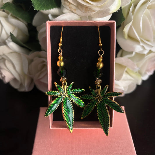 A pair of dangling earrings shaped like green cannabis leaves with gold-tone outlines and small gold beads above, displayed in a pink jewelry box with white roses in the background.