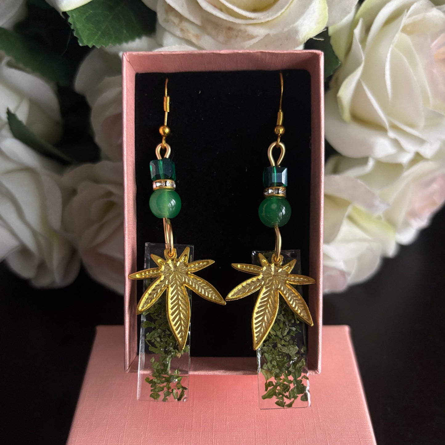 A pair of dangling earrings displayed in an open pink jewelry box. Each earring has a gold-tone hook, a green bead, a green cylindrical spacer, and a gold-tone leaf-shaped charm above a clear square section filled with green dried herbs.
