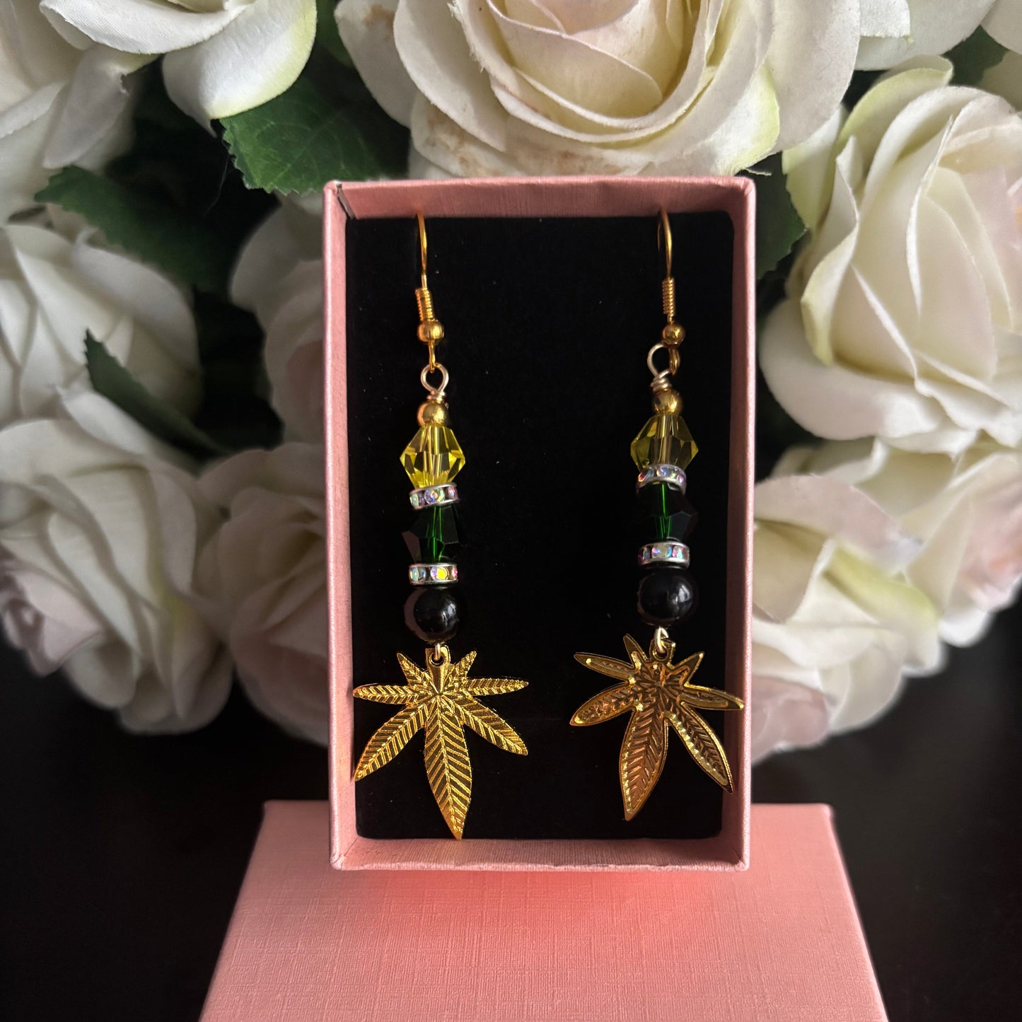 A pair of gold-tone dangling earrings with leaf-shaped charms and green bead accents, displayed in an open pink jewelry box with white roses in the background.