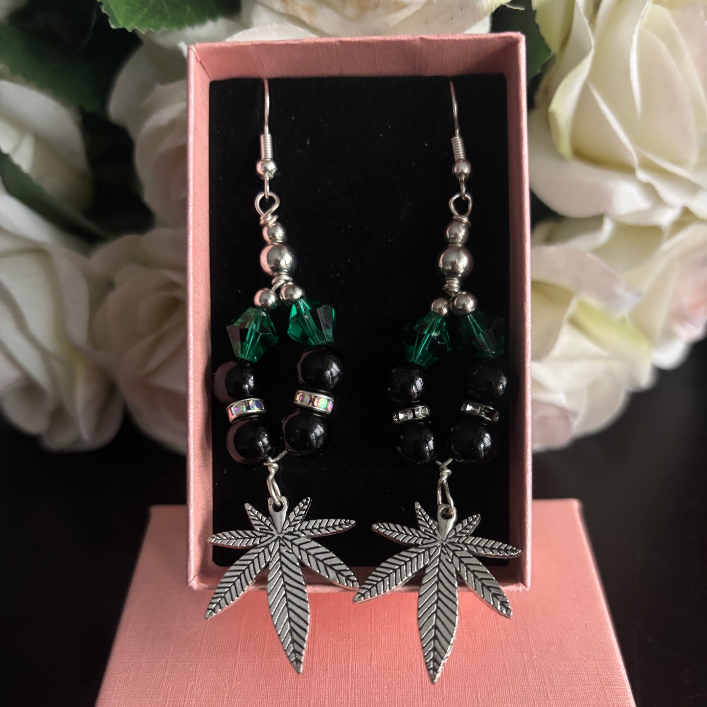 A pair of dangling earrings displayed in a pink jewelry box. The earrings feature silver-tone metal hooks and leaf-shaped charms, with black beads and green faceted beads in the upper section.